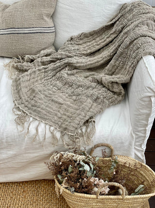 Textured blanket draped over a sofa with a woven basket containing plants on a wooden floor.